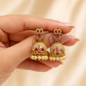 Op de afbeelding: Een paar goudkleurige jhumka-oorbellen met een klokvormig ontwerp ingelegd met heldere kristallen. Elke oorbel is geaccentueerd met een centrale ovaal geslepen roze steen, kleine groene steendetails en een rij bungelende crèmekleurige kralen.
