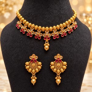 Op de afbeelding: Een goudkleurige ketting en bijpassende oorbellenset met een ketting van getextureerde kralen en bloemvormige motieven. Elke bloem is versierd met donkerroze stenen en heldere kristallen. Een kleine gouden kraal hangt in het midden van de ketting.