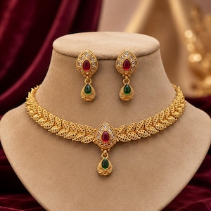 May include: A gold-toned necklace and earring set featuring a leaf-patterned chain. The necklace has a central pendant with a red teardrop stone and a dangling green teardrop stone. The matching earrings feature a red stone and a green teardrop.