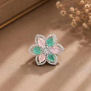 May include: A flower-shaped ring featuring alternating pear-cut pink and green gemstones arranged in a circular pattern. The petals are outlined with small clear stones, and a cluster of clear stones sits at the centre of the floral design.