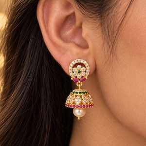 May include: A gold-toned jhumka earring featuring a circular stud with a central green stone surrounded by clear crystals and two pink stones. The bell-shaped drop is adorned with green and pink stones, clear crystals, and a single white pearl.
