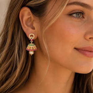 Op de afbeelding: Een goudkleurige jhumka-oorbel met een ronde knop bezet met heldere stenen, een klokvormige hanger versierd met rijen groene, heldere en roze stenen, en een enkele crèmekleurige parel die onderaan het midden van de klokvorm hangt.