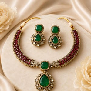 May include: A jewellery set consisting of a necklace and matching earrings. The necklace features a central teardrop-shaped green stone, a square green stone, and bands of deep red stones set in gold-tone metal with small clear accent stones.
