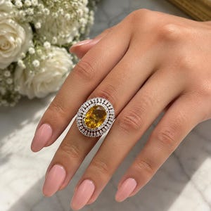 Puede incluir: Un anillo grande, de forma ovalada, con una piedra preciosa amarilla en el centro, rodeada por dos filas de piedras más pequeñas y transparentes. El anillo está engarzado en una banda plateada. La mano que lleva el anillo tiene esmalte de uñas rosa y está cerca de un ramo de flores blancas.