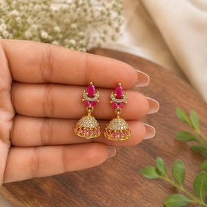 May include: A pair of gold-toned jhumka earrings featuring a teardrop-shaped pink stone at the top, accented by a crescent of small white crystals. The bell-shaped base is adorned with a band of white crystals and a row of small pink stones.
