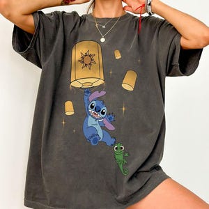 May include: A charcoal gray short-sleeve t-shirt featuring a graphic print of a blue alien character hanging from a yellow floating lantern, accompanied by a small green lizard character and several smaller yellow lanterns against a dark background.