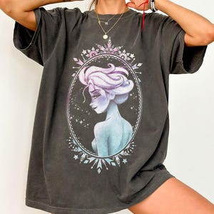 May include: Dark gray t-shirt featuring a portrait of a woman with light purple hair and blue skin, framed in an oval with a decorative border. The design is a stylized illustration with a fantasy theme, printed on a casual, oversized tee.
