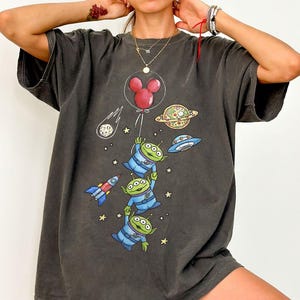 May include: Dark gray t-shirt featuring a graphic of the Toy Story aliens holding a Mickey Mouse balloon, with a rocket ship, planets, and a UFO. The design is in shades of blue, green, red, and yellow. The shirt has short sleeves and a relaxed fit.