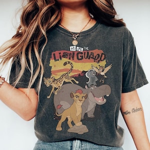 Puede incluir: Camiseta gris oscuro con un gráfico colorido de personajes de la serie animada The Lion Guard. El texto "WE ARE THE LION GUARD" está impreso encima de los personajes. La camiseta tiene un aspecto vintage y desgastado.