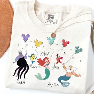 Puede incluir: Camiseta color crema con personajes de La Sirenita. El dise&ntilde;o incluye a &Uacute;rsula, Flounder, Ariel, Sebasti&aacute;n y el Rey Trit&oacute;n, cada uno sosteniendo un globo con forma de Mickey Mouse. La camiseta tiene la etiqueta de la marca Comfort Colors.