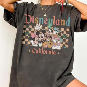 May include: Dark gray t-shirt featuring the word "Disneyland" in colorful text above a graphic of Mickey, Minnie, Goofy, Donald, and Pluto. The shirt also includes the text "Est. 1955 California" with a checkered background.