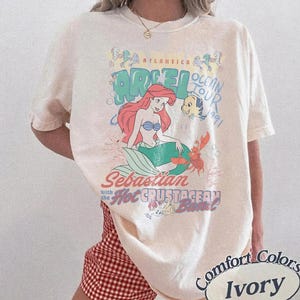 K&ouml;nnte beinhalten: Elfenbeinfarbenes Comfort Colors T-Shirt mit einer Grafik von Arielle, der Meerjungfrau. Das Design zeigt Arielle, Sebastian, Fabius und den Text "Ocean Tour 1991" und "Sebastian with the Hot Crustacean Band!"