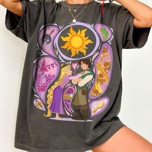 May include: Dark gray t-shirt featuring a colorful graphic of a couple embracing, with a sun, castle, and other elements. The design is framed by a purple border. The shirt has short sleeves and a relaxed fit.