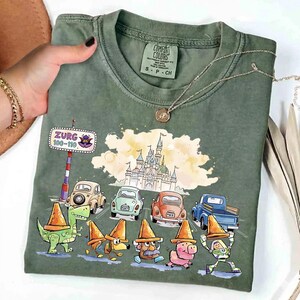 May include: Green Comfort Colors t-shirt featuring a colorful graphic of cartoon characters and cars on a road, with a castle in the background. The shirt has the text "Zurg 100-110" on a road sign. The shirt is size S-P-CH.