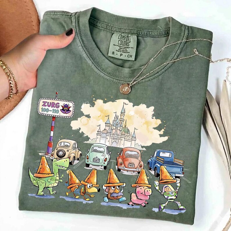 May include: Green Comfort Colors t-shirt featuring a colorful graphic of cartoon characters and cars on a road, with a castle in the background. The shirt has the text "Zurg 100-110" on a road sign. The shirt is size S-P-CH.