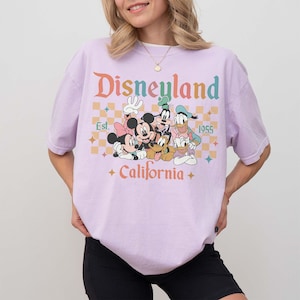 May include: A light purple t-shirt featuring the word "Disneyland" in orange and teal lettering, with a graphic of Mickey Mouse, Minnie Mouse, Goofy, Donald Duck, and Pluto. The shirt also includes the text "Est. 1955" and "California."