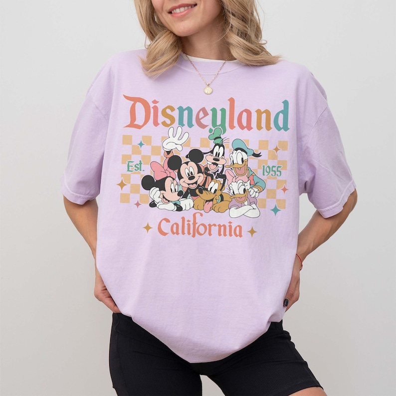 May include: A light purple t-shirt featuring the word "Disneyland" in orange and teal lettering, with a graphic of Mickey Mouse, Minnie Mouse, Goofy, Donald Duck, and Pluto. The shirt also includes the text "Est. 1955" and "California."