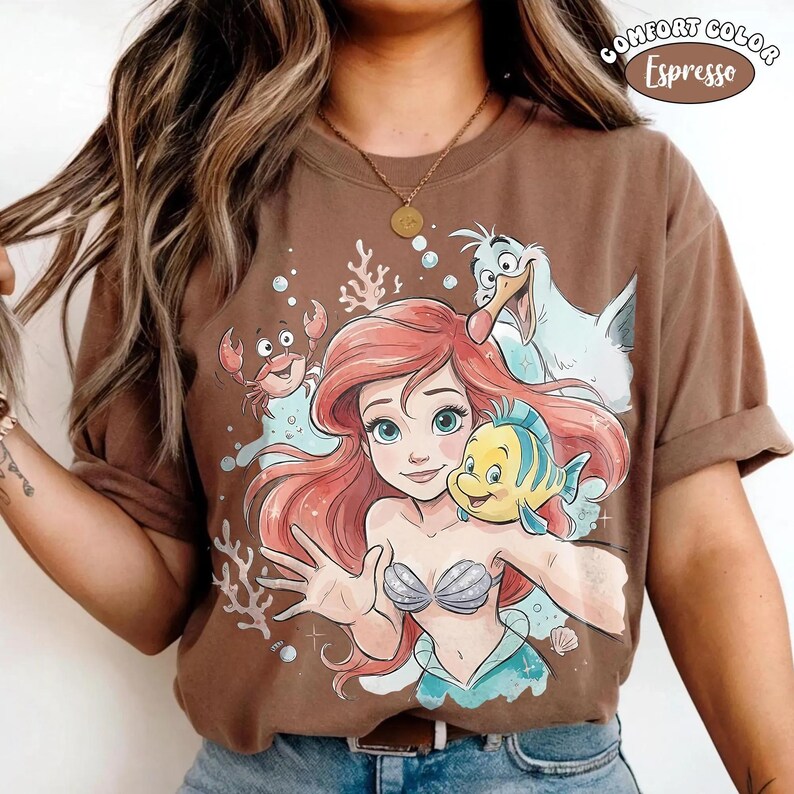 May include: A brown short-sleeve t-shirt featuring a graphic print of a red-haired mermaid, a yellow and blue fish, a small red crab, and a white seagull. The design includes coral and bubbles. Text in the upper right corner reads COMFORT COLOR Espresso.