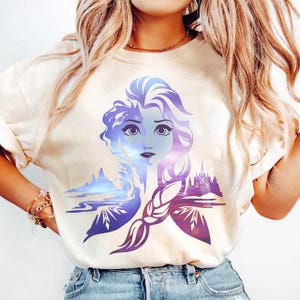 以下が含まれることがあります： クリーム色のTシャツ。青と紫の色合いで女性の顔をスタイリッシュに表現したイラストが描かれています。デザインにはお城や風景の要素が含まれており、三つ編みの髪型です。カジュアルウェアです。