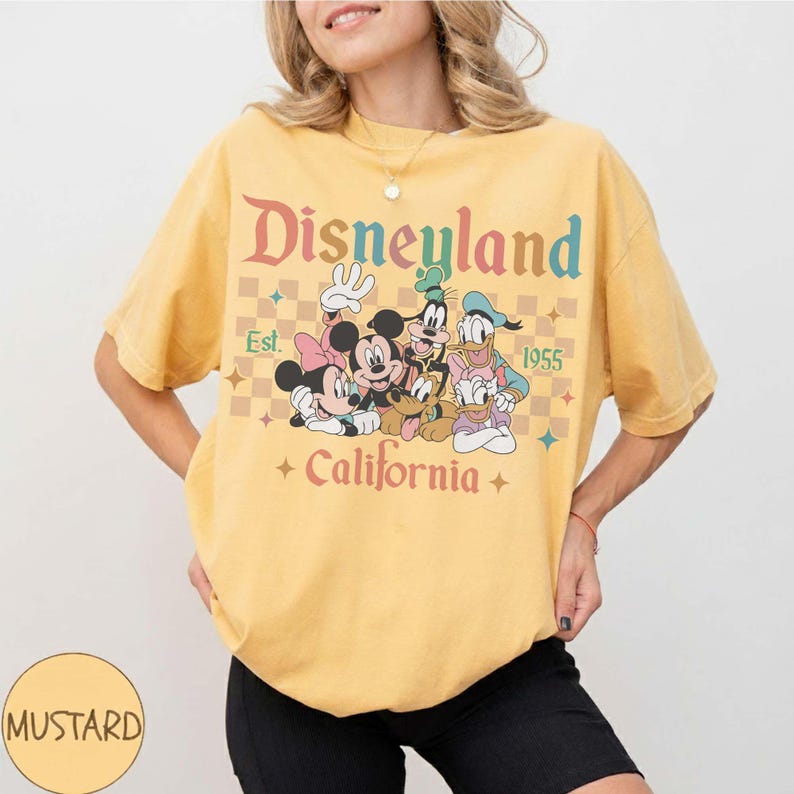 May include: Mustard yellow t-shirt featuring the Disneyland logo with Mickey Mouse, Minnie Mouse, Goofy, Donald Duck, and Daisy Duck. The shirt has the text "Est. 1955" and "California".