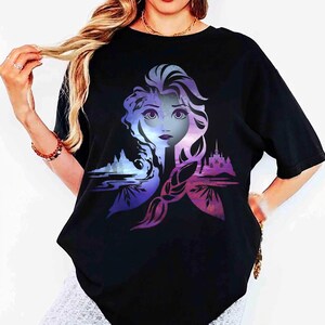以下が含まれることがあります： 紫と青の色合いの女性の顔のグラフィックが特徴の黒いTシャツ。デザインには城と風景の要素が含まれています。シャツは半袖でカジュアルなスタイルです。