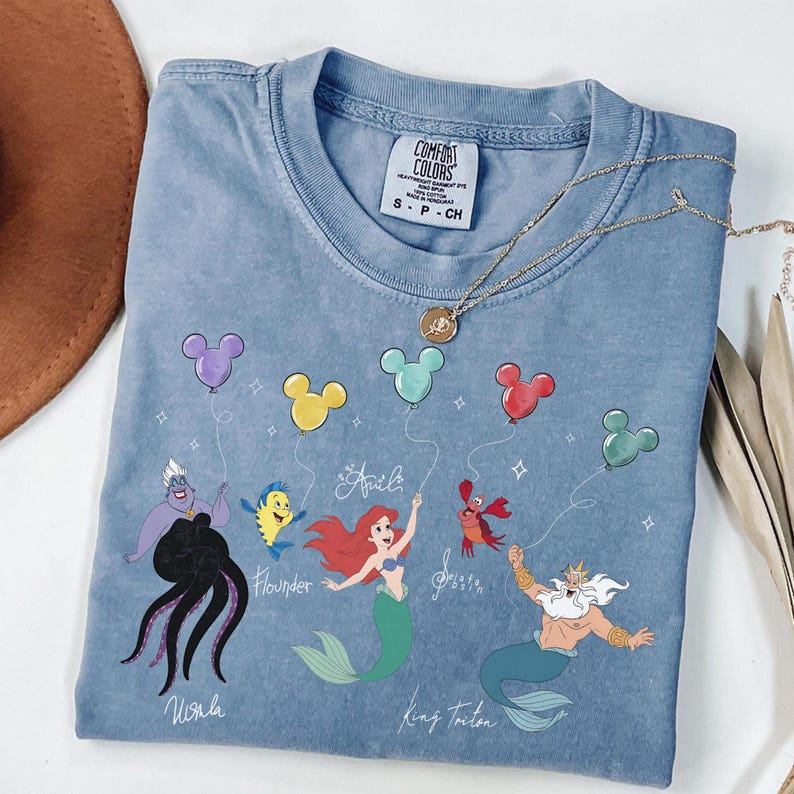 Puede incluir: Una camiseta azul Comfort Colors con un gr&aacute;fico de personajes de La Sirenita, incluyendo a Ariel, Flounder, Sebasti&aacute;n, el Rey Trit&oacute;n y &Uacute;rsula, cada uno sosteniendo un globo de Mickey Mouse. Un collar de oro descansa sobre la camisa.