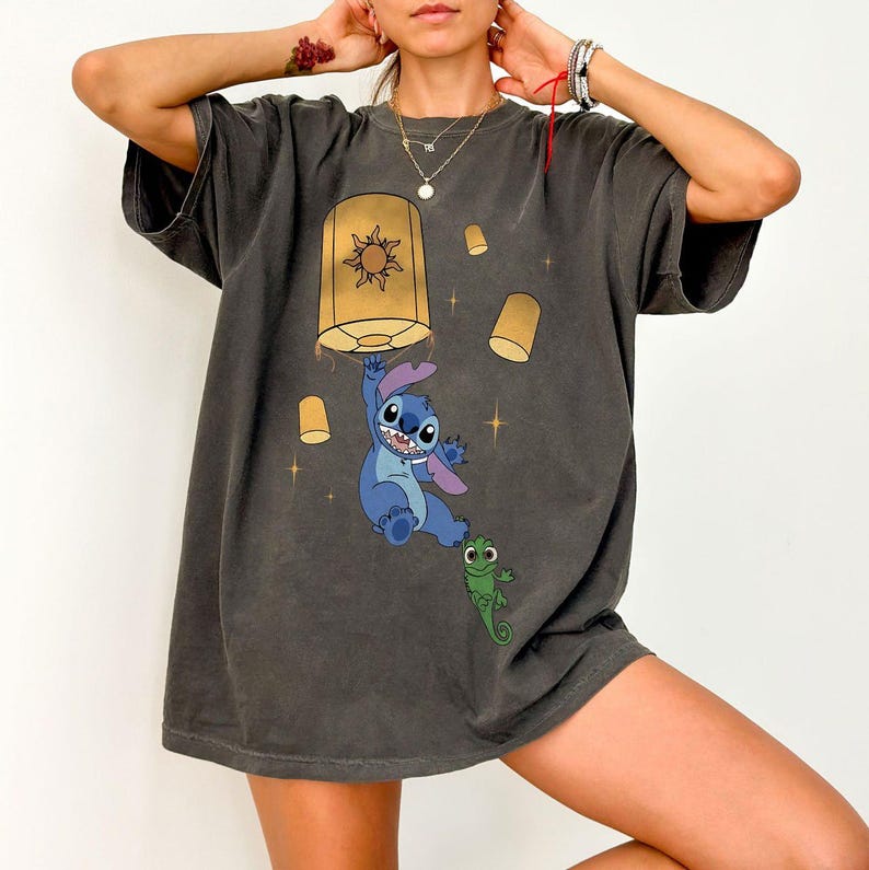 May include: A charcoal gray short-sleeve t-shirt featuring a graphic print of a blue alien character hanging from a yellow floating lantern, accompanied by a small green lizard character and several smaller yellow lanterns against a dark background.
