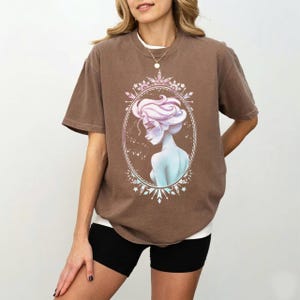 May include: A brown t-shirt featuring a portrait of a woman with light blue skin and pink hair, framed in an oval with a snowflake design. The woman is wearing a white undershirt and black shorts.