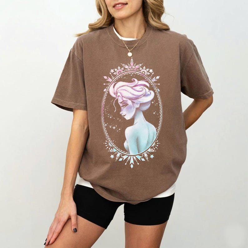 May include: A brown t-shirt featuring a portrait of a woman with light blue skin and pink hair, framed in an oval with a snowflake design. The woman is wearing a white undershirt and black shorts.