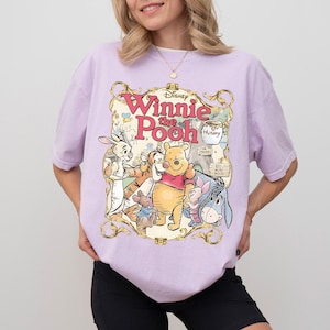 May include: A light purple t-shirt featuring a colorful graphic of Winnie the Pooh and friends. The design includes the text "Winnie the Pooh" in a decorative frame, with characters like Pooh Bear, Tigger, and Eeyore.