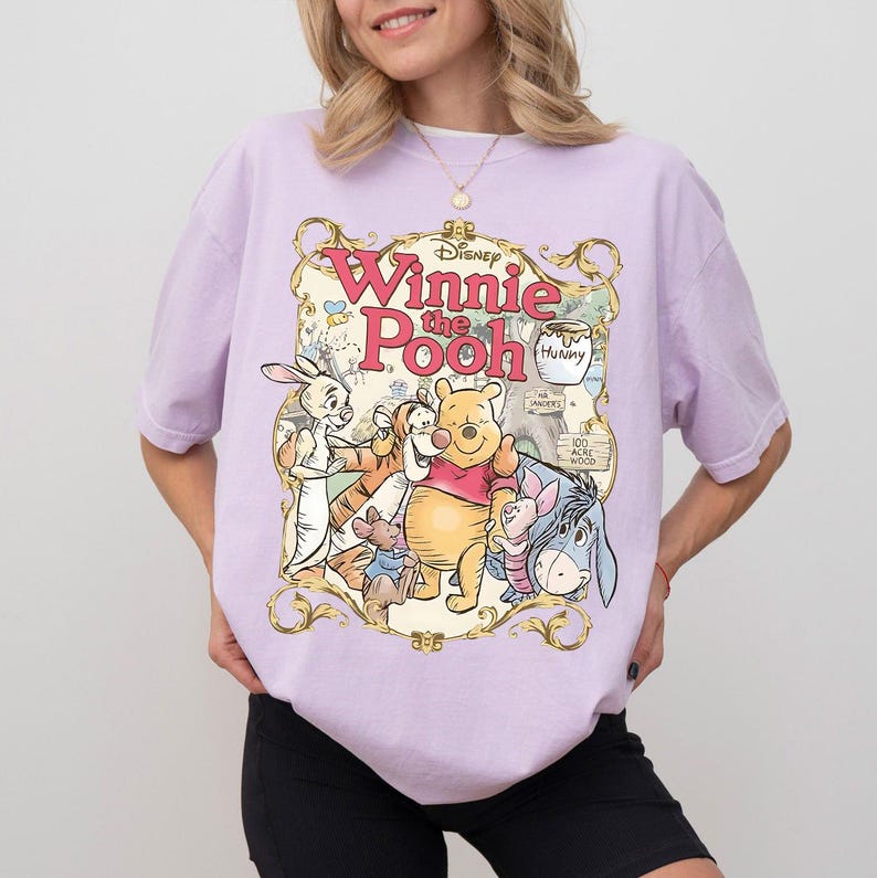 May include: A light purple t-shirt featuring a colorful graphic of Winnie the Pooh and friends. The design includes the text "Winnie the Pooh" in a decorative frame, with characters like Pooh Bear, Tigger, and Eeyore.