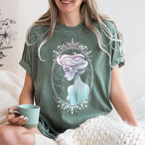 May include: Green t-shirt featuring a watercolor illustration of a woman with light purple hair and blue skin, framed in an oval with stars and snowflakes. The shirt is a solid green color.
