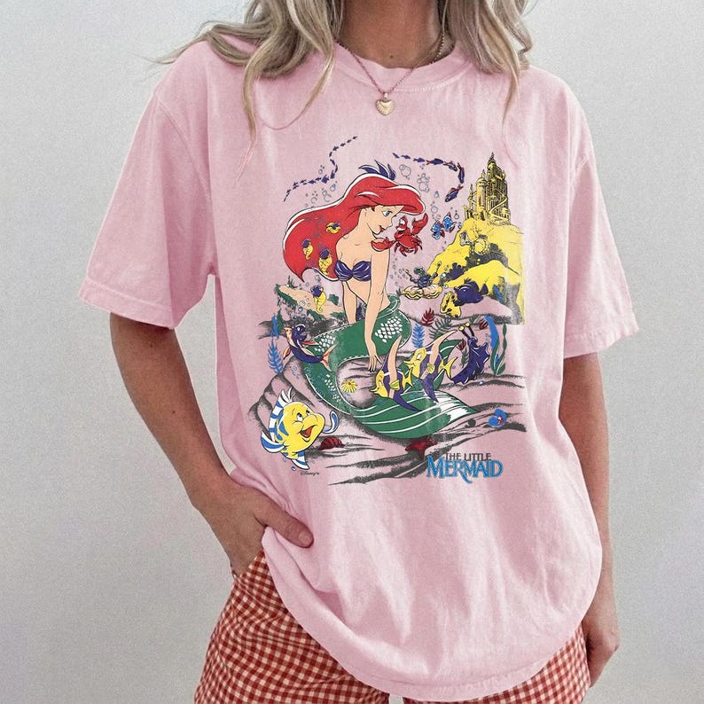 K&ouml;nnte beinhalten: Hellrosa T-Shirt mit einem farbenfrohen Aufdruck von Arielle, der Meerjungfrau. Die Illustration zeigt Arielle mit roten Haaren und einem gr&uuml;nen Schwanz, sowie den Text "THE LITTLE MERMAID" in Blau.
