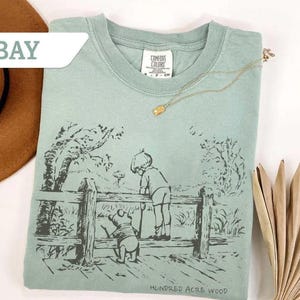 May include: A light green t-shirt featuring a black and white illustration of a boy and a bear looking over a wooden fence. The text "Hundred Acre Wood" is printed at the bottom. A gold necklace is draped across the shirt.
