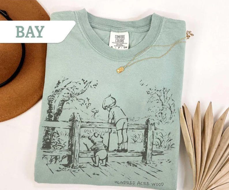 May include: A light green t-shirt featuring a black and white illustration of a boy and a bear looking over a wooden fence. The text "Hundred Acre Wood" is printed at the bottom. A gold necklace is draped across the shirt.