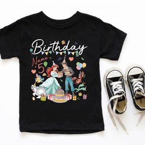 Pode incluir: T-shirt preto de manga curta com um gr&aacute;fico colorido com tema de anivers&aacute;rio. O design inclui a palavra "Birthday" em escrita cursiva, o n&uacute;mero "5" e personagens de desenhos animados. Um par de t&ecirc;nis de lona pretos com cadar&ccedil;os brancos est&aacute; ao lado da camisa.