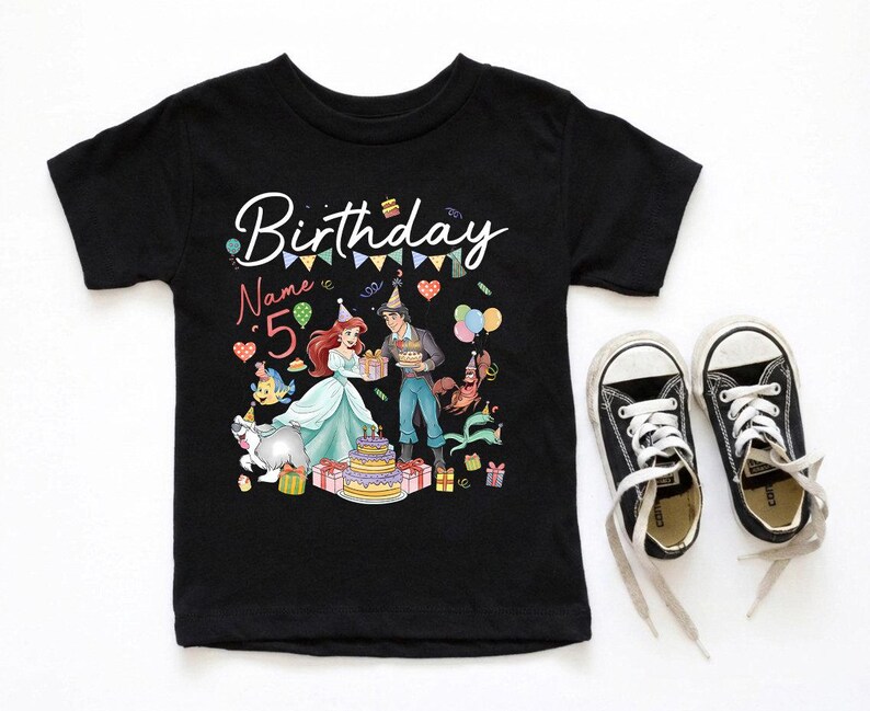 Pode incluir: T-shirt preto de manga curta com um gr&aacute;fico colorido com tema de anivers&aacute;rio. O design inclui a palavra "Birthday" em escrita cursiva, o n&uacute;mero "5" e personagens de desenhos animados. Um par de t&ecirc;nis de lona pretos com cadar&ccedil;os brancos est&aacute; ao lado da camisa.