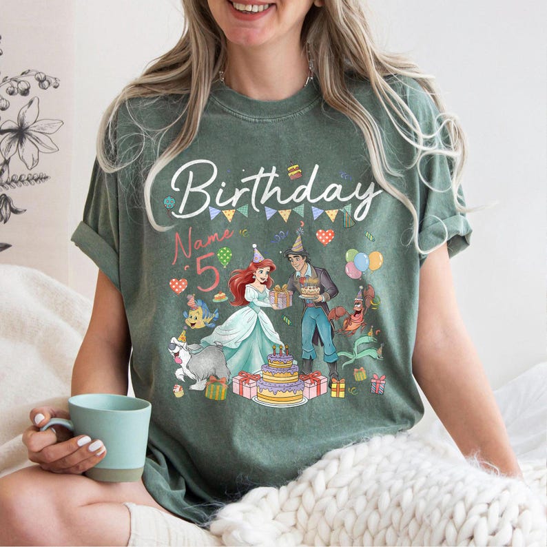 Pode incluir: Camiseta verde com um gr&aacute;fico de anivers&aacute;rio com Ariel e Pr&iacute;ncipe Eric, um bolo e o n&uacute;mero 5. A palavra "Birthday" est&aacute; escrita em it&aacute;lico acima do gr&aacute;fico. A camisa tem mangas enroladas.