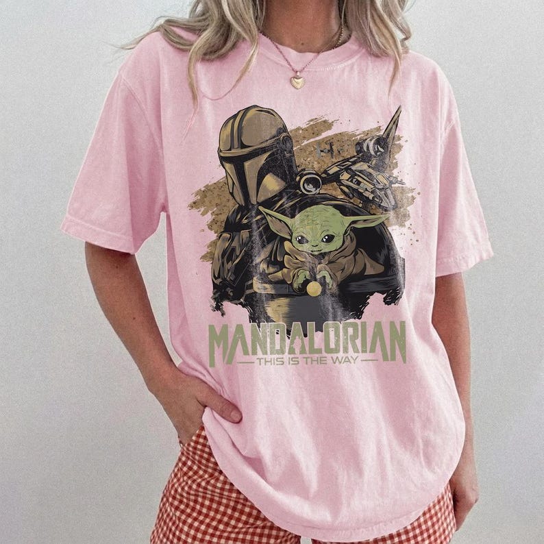 May include: Pink t-shirt featuring a graphic of The Mandalorian and Grogu from the Star Wars series. The design includes the text "MANDALORIAN - THIS IS THE WAY" in a green and white font. The shirt is a light pink color.