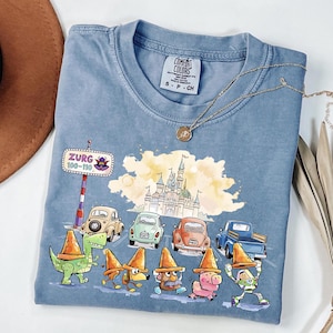 May include: A blue Comfort Colors t-shirt featuring a colorful graphic of cartoon characters, cars, and a castle. The graphic includes the text "Zurg 100-110". A gold necklace and a brown hat are also visible.