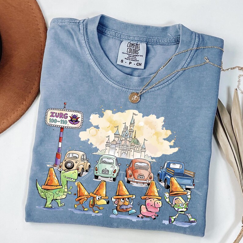 May include: A blue Comfort Colors t-shirt featuring a colorful graphic of cartoon characters, cars, and a castle. The graphic includes the text "Zurg 100-110". A gold necklace and a brown hat are also visible.