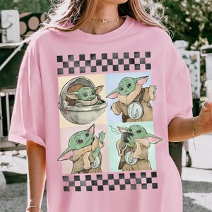 Puede incluir: Camiseta rosa claro con un dise&ntilde;o de cuatro paneles de un personaje alien&iacute;gena verde. El dise&ntilde;o est&aacute; enmarcado por patrones de cuadros blancos y negros. El personaje se muestra en una c&aacute;psula, sosteniendo una taza y gesticulando.