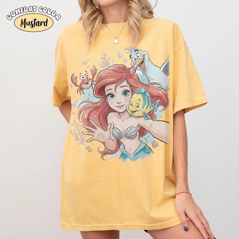 May include: A mustard yellow short-sleeve t-shirt featuring a graphic print of a red-haired mermaid, a yellow and blue fish, a red crab, and a white seagull. The text COMFORT COLOR MUSTARD is printed in the upper left corner of the shirt design.