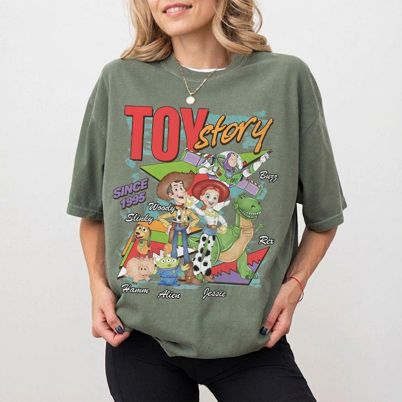 K&ouml;nnte beinhalten: Ein olivgr&uuml;nes T-Shirt mit den Worten "Toy Story" in Rot und Gelb sowie Illustrationen von Woody, Buzz, Jessie und Rex. Das Shirt enth&auml;lt auch den Text "Since 1995" und die Namen der Charaktere. Das T-Shirt hat eine lockere Passform.