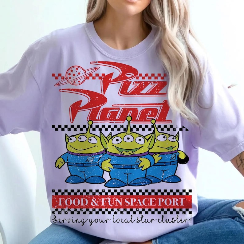 May include: A light purple t-shirt featuring a red graphic with the text Pizza Planet. The design includes three green three-eyed aliens in blue space suits, a planet icon, and a checkered border. Below is the text FOOD & FUN SPACE PORT.