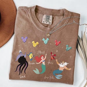 Puede incluir: Una camiseta marr&oacute;n Comfort Colors con un gr&aacute;fico de los personajes de La Sirenita de Disney sosteniendo globos con forma de Mickey Mouse. Los personajes incluyen a Ariel, Flounder, Sebasti&aacute;n, &Uacute;rsula y el Rey Trit&oacute;n. Un collar plateado con un colgante est&aacute; sobre la camiseta.