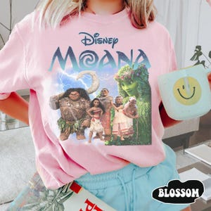 Pu&ograve; includere: T-shirt rosa chiaro con una grafica dei personaggi del film Disney Moana, con il titolo in blu. La maglietta &egrave; abbinata a pantaloncini blu. Nella foto &egrave; tenuta una tazza con una faccina sorridente.