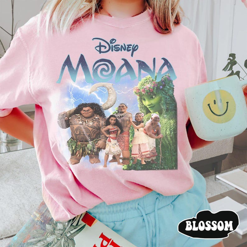 Pu&ograve; includere: T-shirt rosa chiaro con una grafica dei personaggi del film Disney Moana, con il titolo in blu. La maglietta &egrave; abbinata a pantaloncini blu. Nella foto &egrave; tenuta una tazza con una faccina sorridente.