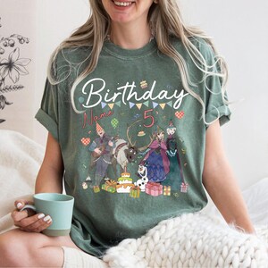 May include: Green t-shirt with a birthday theme. The shirt features the word "Birthday" in white script, the number "5", and cartoon characters. Colorful confetti and gift boxes are also visible on the shirt.