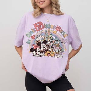 May include: A light purple t-shirt featuring a colorful graphic of Disney characters, including Mickey Mouse, Minnie Mouse, Donald Duck, Goofy, and Pluto, with the text "Walt Disney World" above them. The design includes a rainbow and a castle.